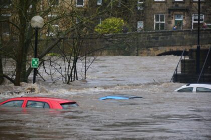 Three cars partially submerged in a flooded river near a stone bridge