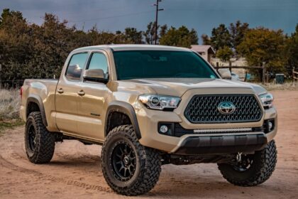 A tan Toyota Tacoma pickup truck on a dirt road with black wheels